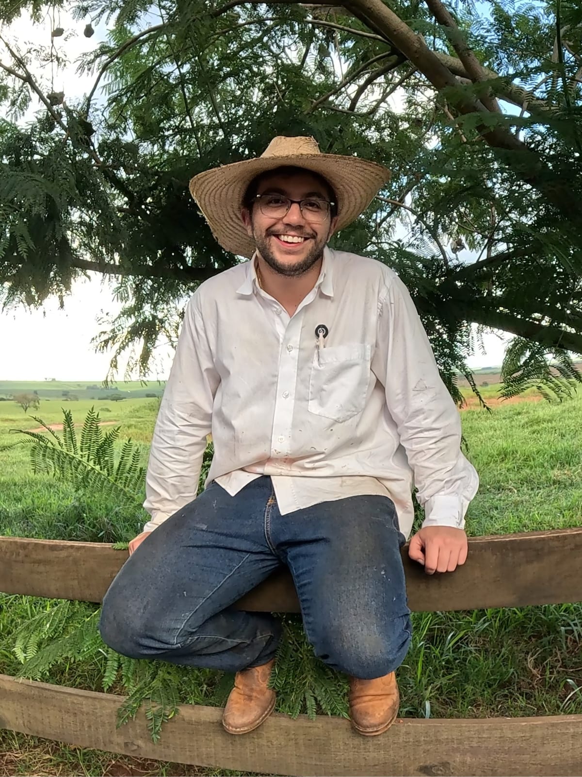 Pedro, de chapéu, sentado na cerca da fazenda com o pasto ao fundo